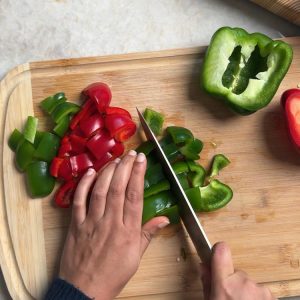 Hands cutting red and green bell peppers into square chunks on a bamboo cutting board