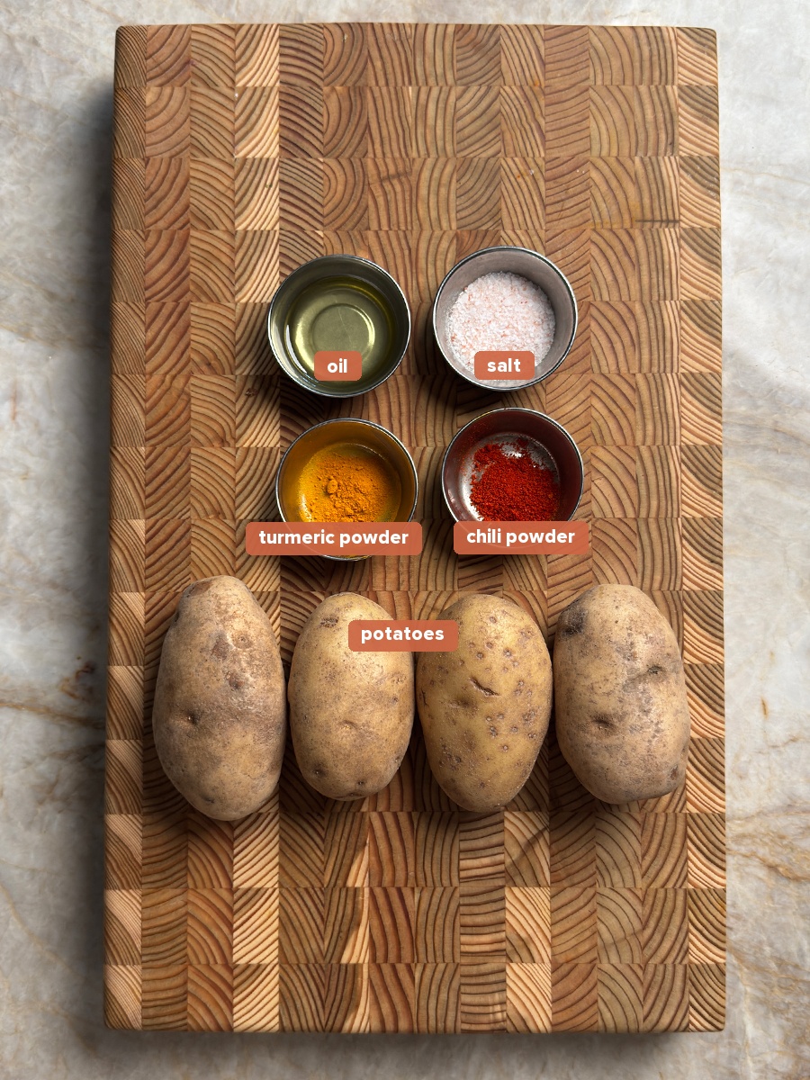 Potatoes, oil, salt, turmeric, and chili powder arranged on a wooden cutting board