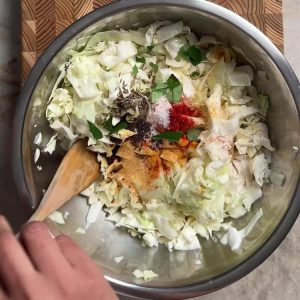 Chopped cabbage in a metal bowl with turmeric, chili powder, cumin seeds, mustard seeds, and curry leaves being mixed in
