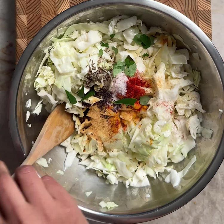 Chopped cabbage in a metal bowl with turmeric, chili powder, cumin seeds, mustard seeds, and curry leaves being mixed in