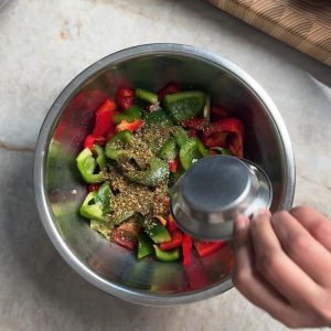Adding Mediterranean herb blend from a small cup to oiled peppers in a mixing bowl