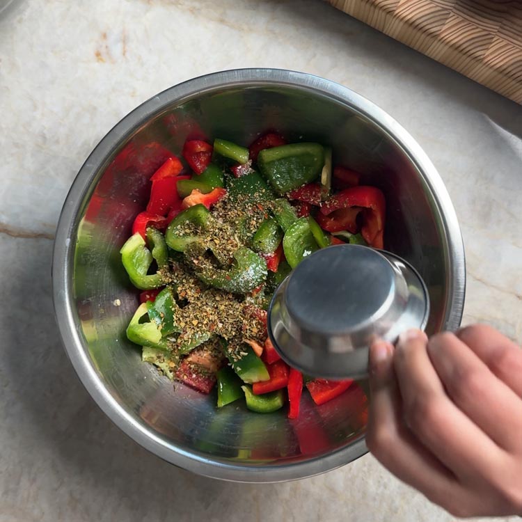 Adding Mediterranean herb blend from a small cup to oiled peppers in a mixing bowl
