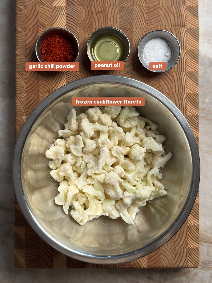 Frozen cauliflower florets in a mixing bowl with small bowls of garlic chili powder, peanut oil, and salt on a wooden cutting board