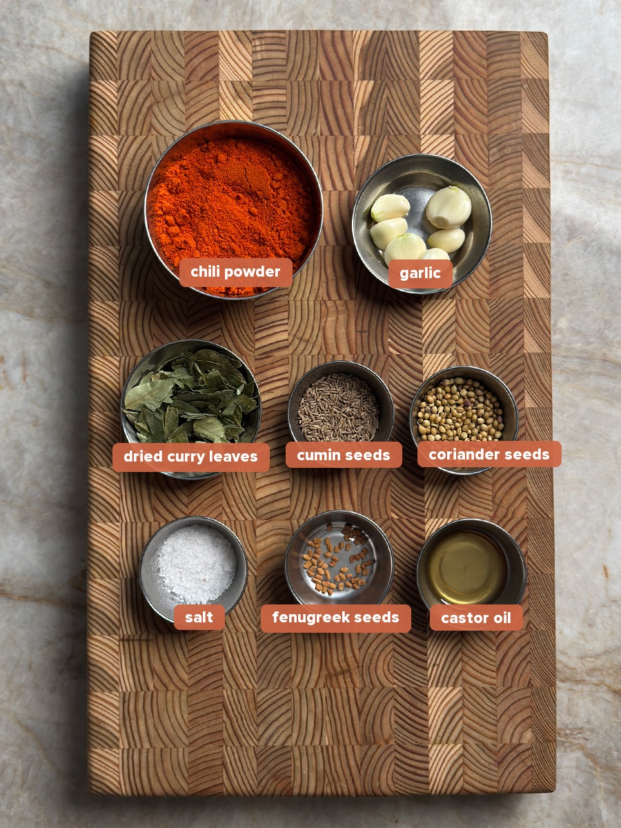 Eight steel bowls of whole spices and garlic cloves arranged on a wooden end grain cutting board: chili powder, curry leaves, cumin seeds, coriander seeds, salt, fenugreek seeds and castor oil