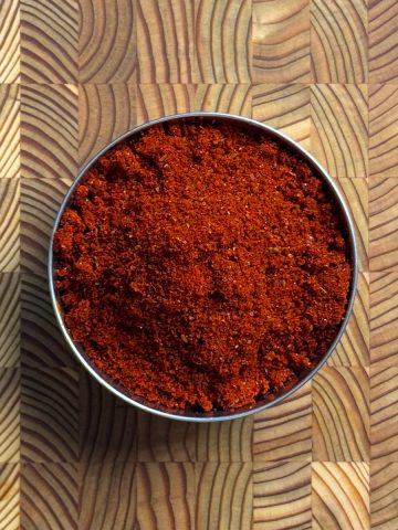 Deep red garlic chili powder heaped in a round steel tin on a wooden cutting board, fine grained texture