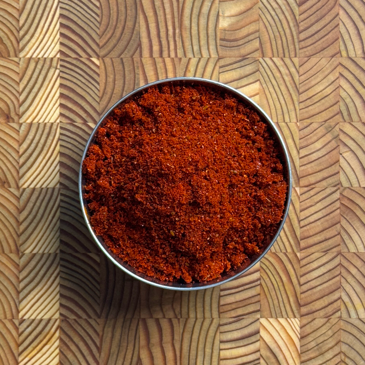 Deep red garlic chili powder heaped in a round steel tin on a wooden cutting board, fine grained texture