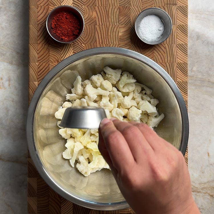 Hand pouring peanut oil from a small cup onto frozen cauliflower florets in a stainless steel mixing bowl