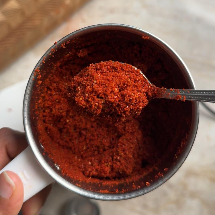 A hand holds a spoon scooping coarsely ground red garlic podi from the grinder jar