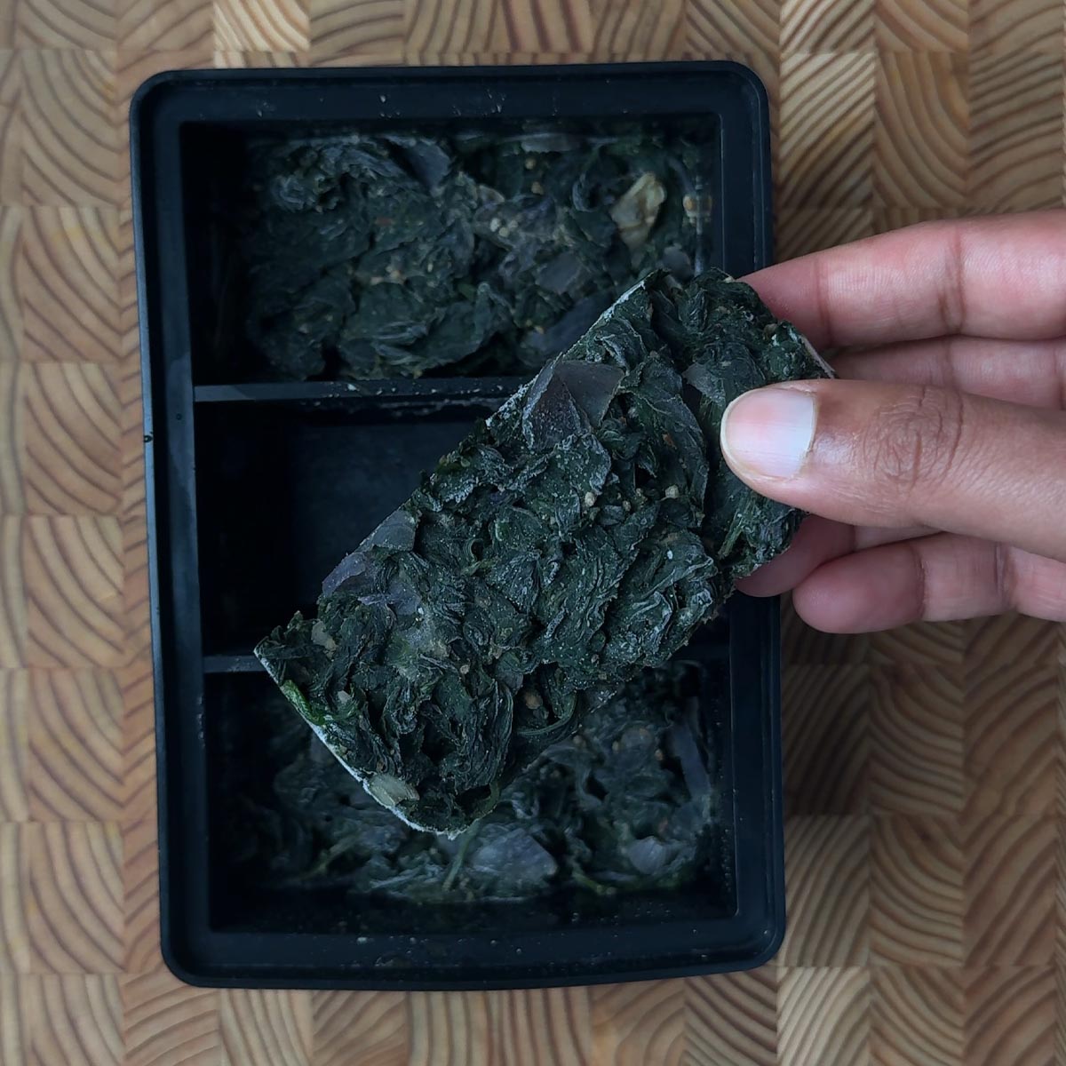 Hand holding a solid frozen spinach fry cube above a black freezer tray