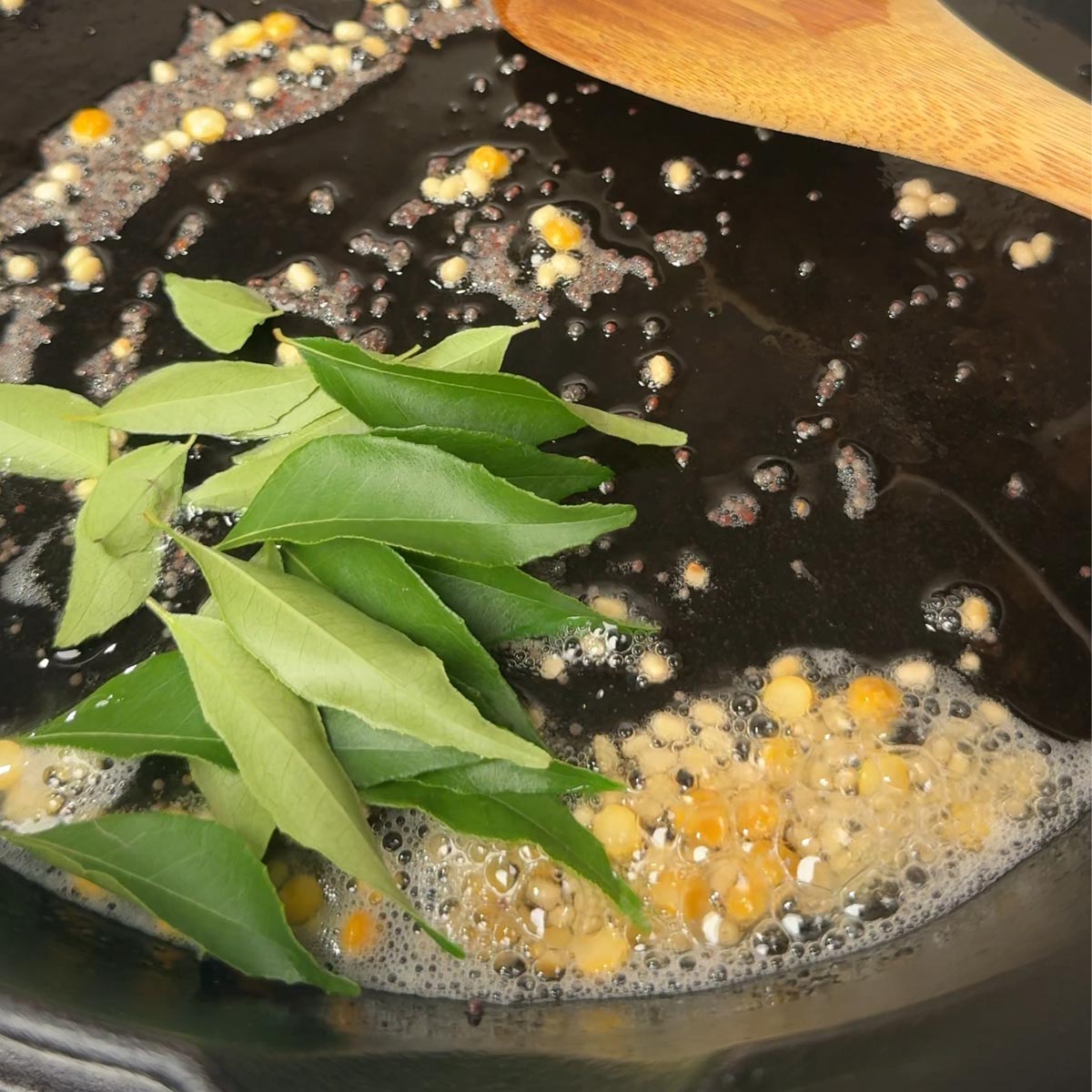 Fresh curry leaves added to hot peanut oil with mustard seeds and golden dal in a black pan