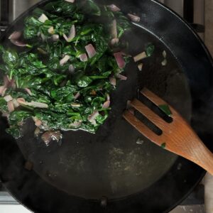 Spinach and onions cooking down in a black pan on the stovetop, steam rising