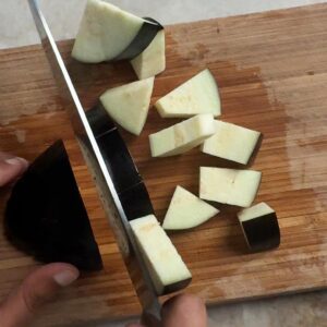 Hands cutting a whole eggplant into cubes on a wooden cutting board with a knife