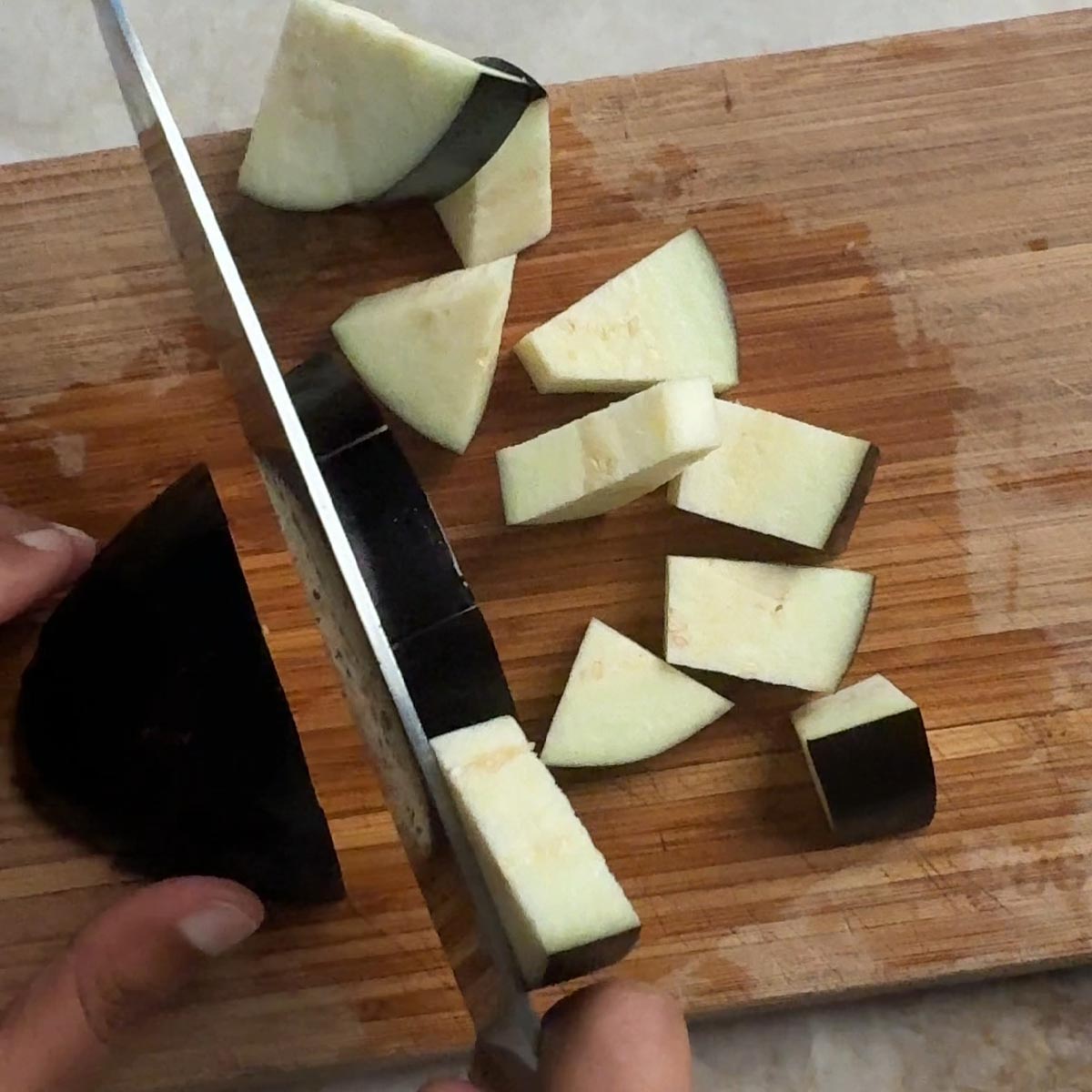 Hands cutting a whole eggplant into cubes on a wooden cutting board with a knife