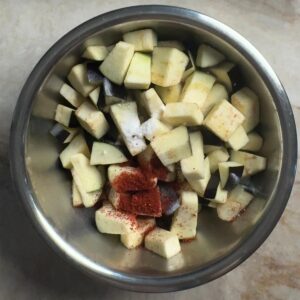Eggplant cubes in a stainless steel mixing bowl with chili powder and salt added before tossing
