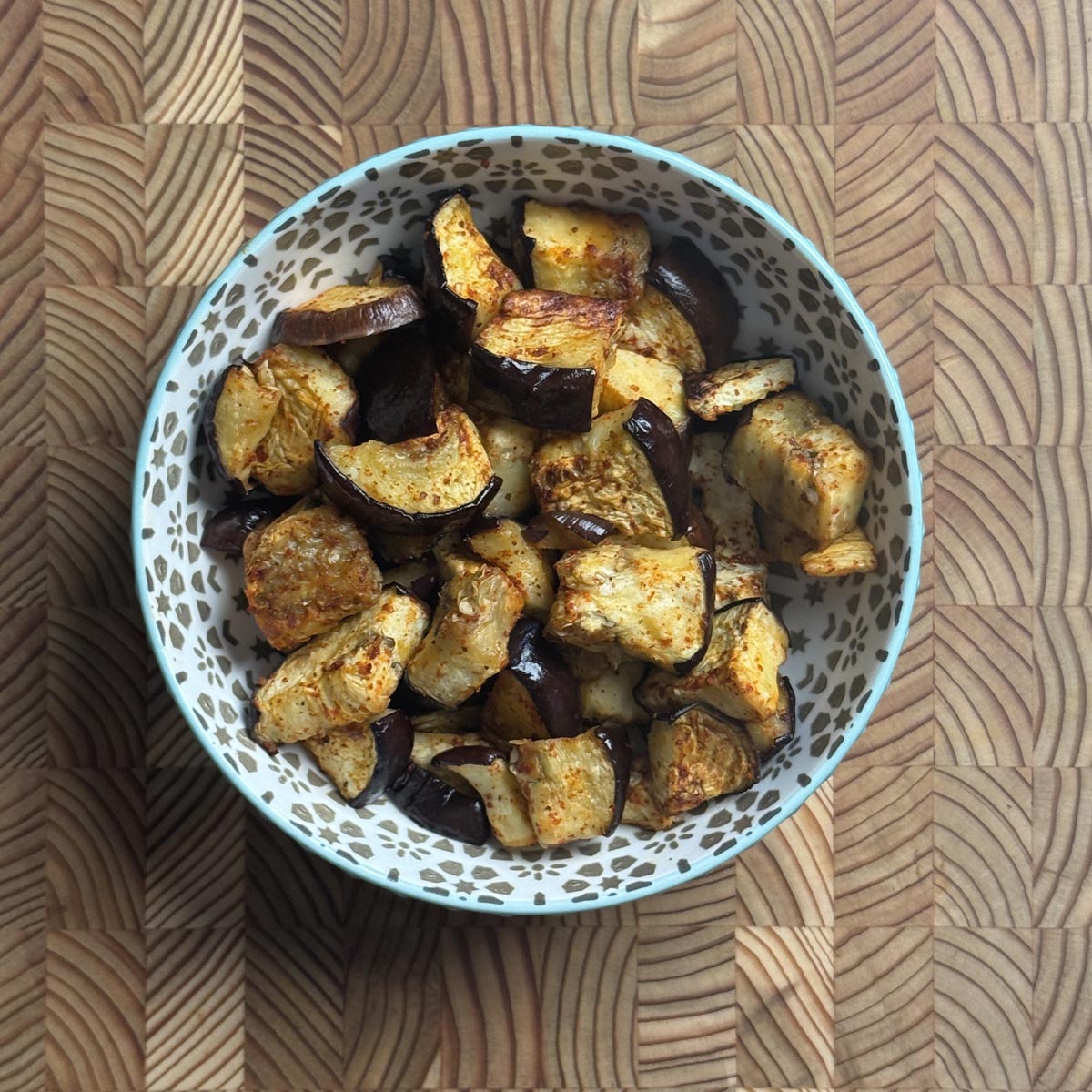 Oven roasted eggplant cubes in a teal patterned bowl on a wooden cutting board, overhead view