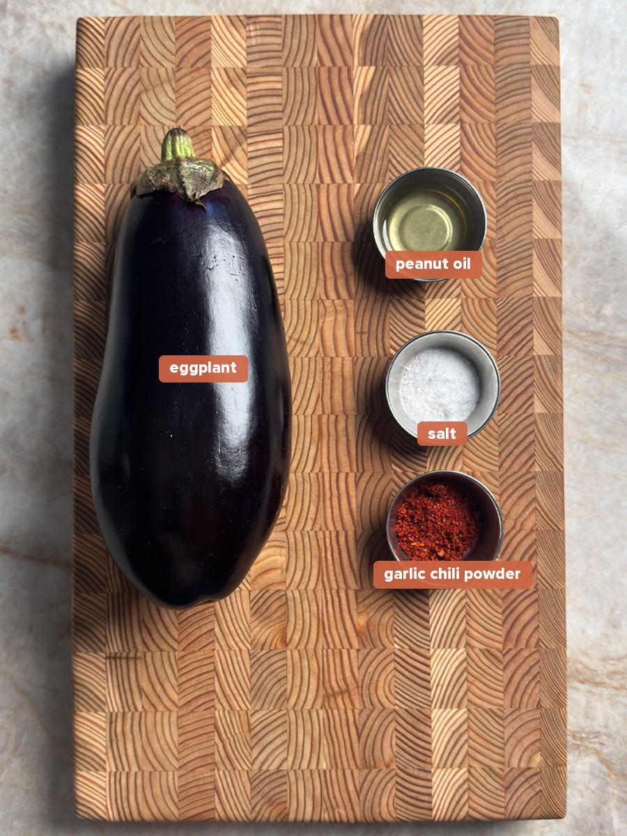 Ingredients for oven roasted eggplant: whole eggplant, peanut oil, salt, and garlic chili powder on a wooden board