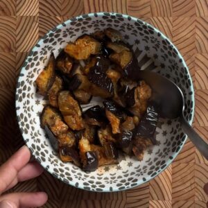 Roasted eggplant in a patterned bowl with a spoon, ready to serve, held over a wooden board