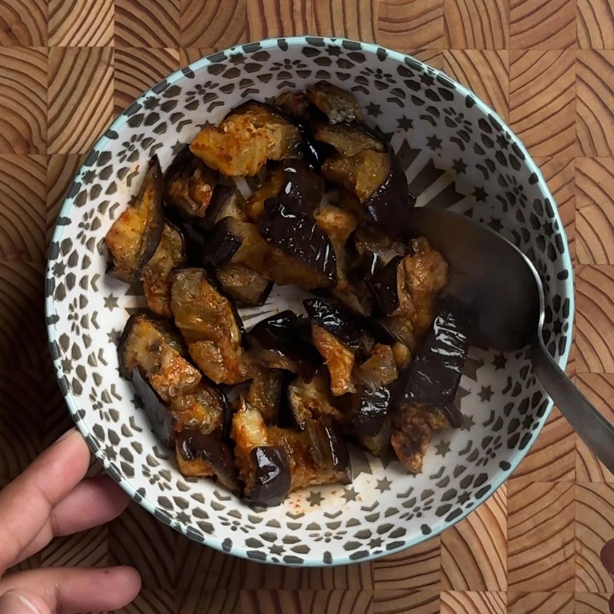 Roasted eggplant in a patterned bowl with a spoon, ready to serve, held over a wooden board