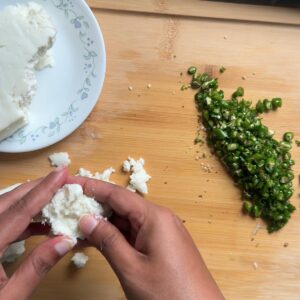 Hands crumbling a block of paneer next to a pile of finely chopped green chilies on a wood board.
