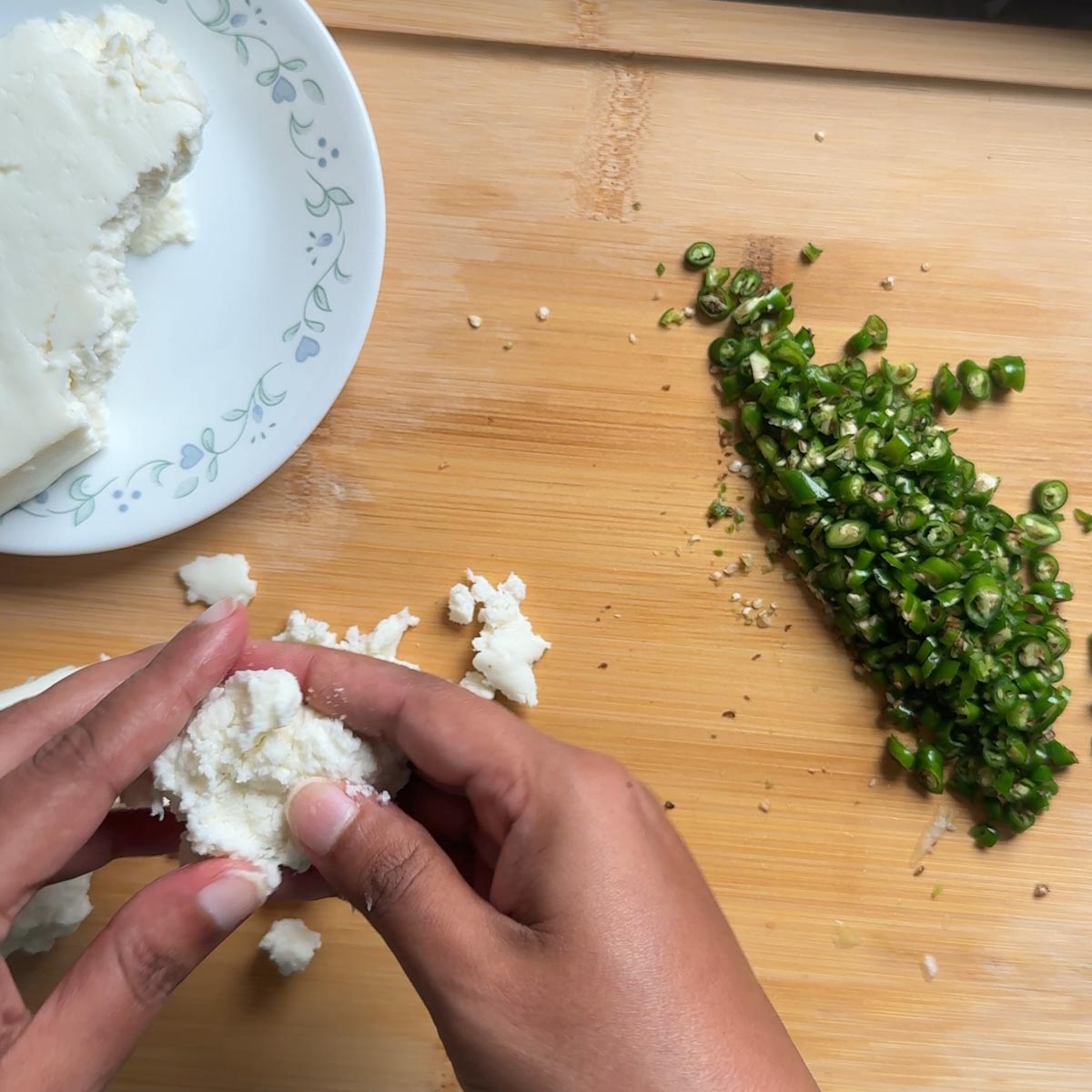 Hands crumbling a block of paneer next to a pile of finely chopped green chilies on a wood board